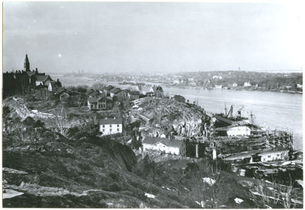 Svartvitt foto av Finnboda varv sett från höjd, med varvsbyggnader och fartyg vid vattnet samt Stockholm i bakgrunden.