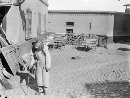 En gammal man med träben och käpp på gården till Stockholms stads allmänna försörjningsinrättning som i folkmun kallas Grubbens.