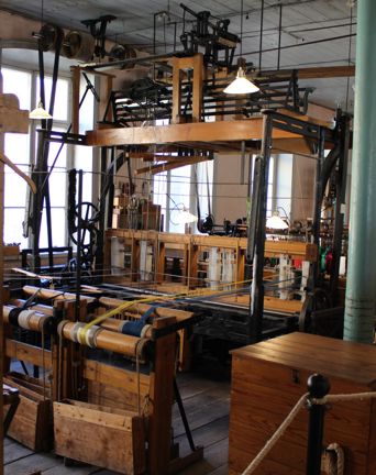 En stor maskin av järn och trä står på ett trägolv. I maskinen syns blåa och gula trådar uppspända som ska vävas till band. Bakom maskinen syns ett stort fönster.