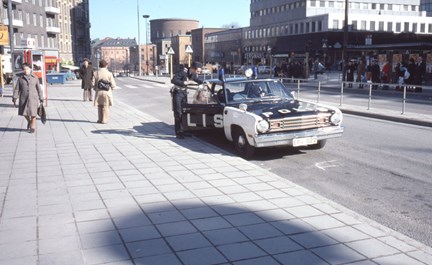 Polisbil, två poliser och passerande människor. Stadsbiblioteket i bakgrunden.