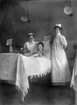 Emilia Gustafsson i en sjukhussäng tillsammans med två uniformerade sjuksystrar. Bilden är arrangerad och visar tre arbetskamrater under en period då Emilia Gustafsson arbetade på Sabbatsbergs sjukhus