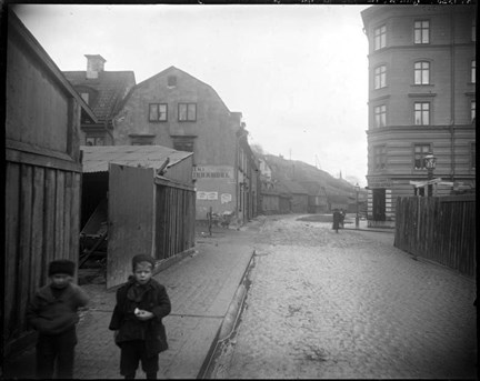 Svartvitt fotografi på en stenlagd gata med trottoar. Plankbebyggelse till vänster och höger om gatan. Två barn i förgrunden till vänster.  I mellangrunden syns stenhus, i bakgrunden ett berg.