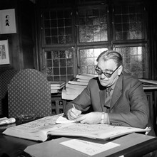 Vilhelm Moberg vid ett skrivbord, läsandes tidningslägg från Svenska Dagbladet