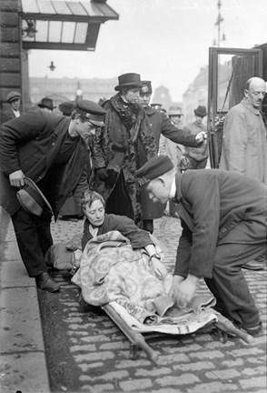 En kvinna på bår tas om hand av ett antal män i uniform. Båren ligger direkt på gatan. Den skadade får hjälp med att rätta till filten hon har över sig.