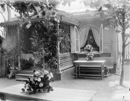 Ett lusthus med en veranda, täckt av växtlighet. På ett bord framför huset ligger en blombukett. Ett fönster står öppet med en gardin och mer blombuketter