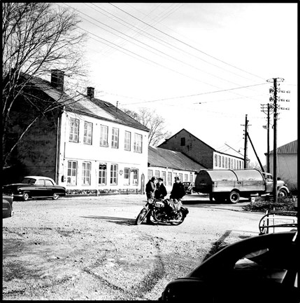 Verkstadsbebyggelse. På planen framför byggnaderna står tre män vid en motorcykel. En gammal tankbil står parkerad i bakgrunden.