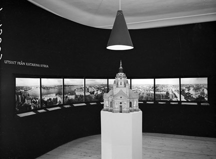 I centrum för bilden står en modell av Katarina kyrka placerad under en lampa. På väggen i bakgrunden syns åtta fotografier som visar olika utsikter från Katarinaberget.
