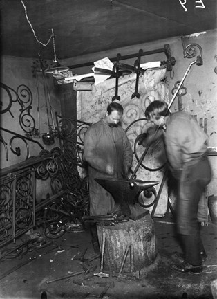 Svart-vit bild med två män i en verkstads-miljö. På golvet står en stubbe med ett städ och flera verktyg. De två männen hjälps åt att smida en lång stång.