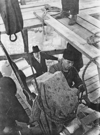 Svartvitt fotografi fotat uppifrån ner över ett arbetslag som lyfter upp en glänsande krona med rep uppför byggställningar. En man i mössa och arbetskläder står i centrum och en man i hatt och rock syns i bakgrunden.