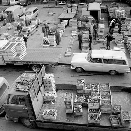 En mängd trälådor med frukt och grönsaker är staplade på varandra. Bilar och mindre lastbilar står parkerade intill. Människor packar upp varor och gör affärer.