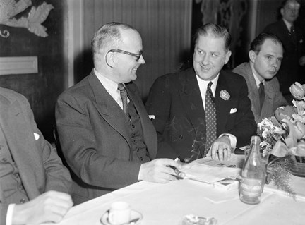 Kostymklädda män vid ett bord med vit duk och blomsterdekoration. Gösta Bergman röker.