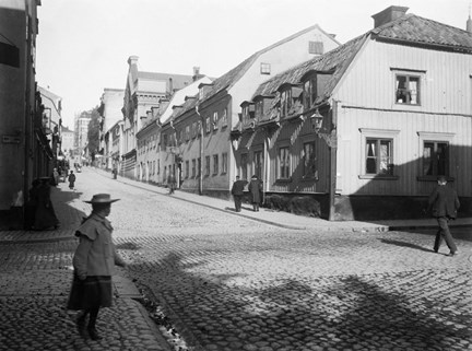 En flicka i 10-årsåldern står i en korsning av två gatstensbelagda gator. Den större gatan kantas av låga hus av trä och sten. Flickan är finklädd och klädena ser ut att vara från sekelskiftet 1900.