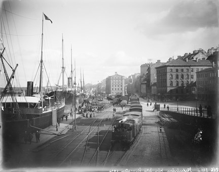Svartvit bild med båtar, tåg och byggnader. I främre delen av bilden syns tågspår och flera vagnar. Till vänster ligger båtar i hamn.
